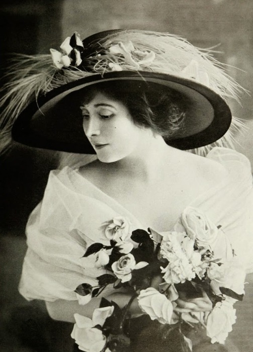 Красоту девушки украшает изысканная шляпа украшенная перьями и нежный букет белых роз.