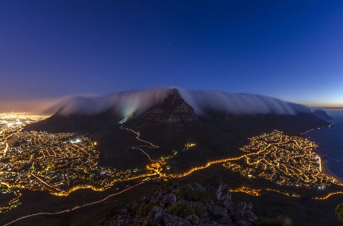 Столовая гора окутана в одеяло из тумана, который медленно сползает на мерцающий в ночи Кейптаун. Фотограф - Brendon Wainwright.